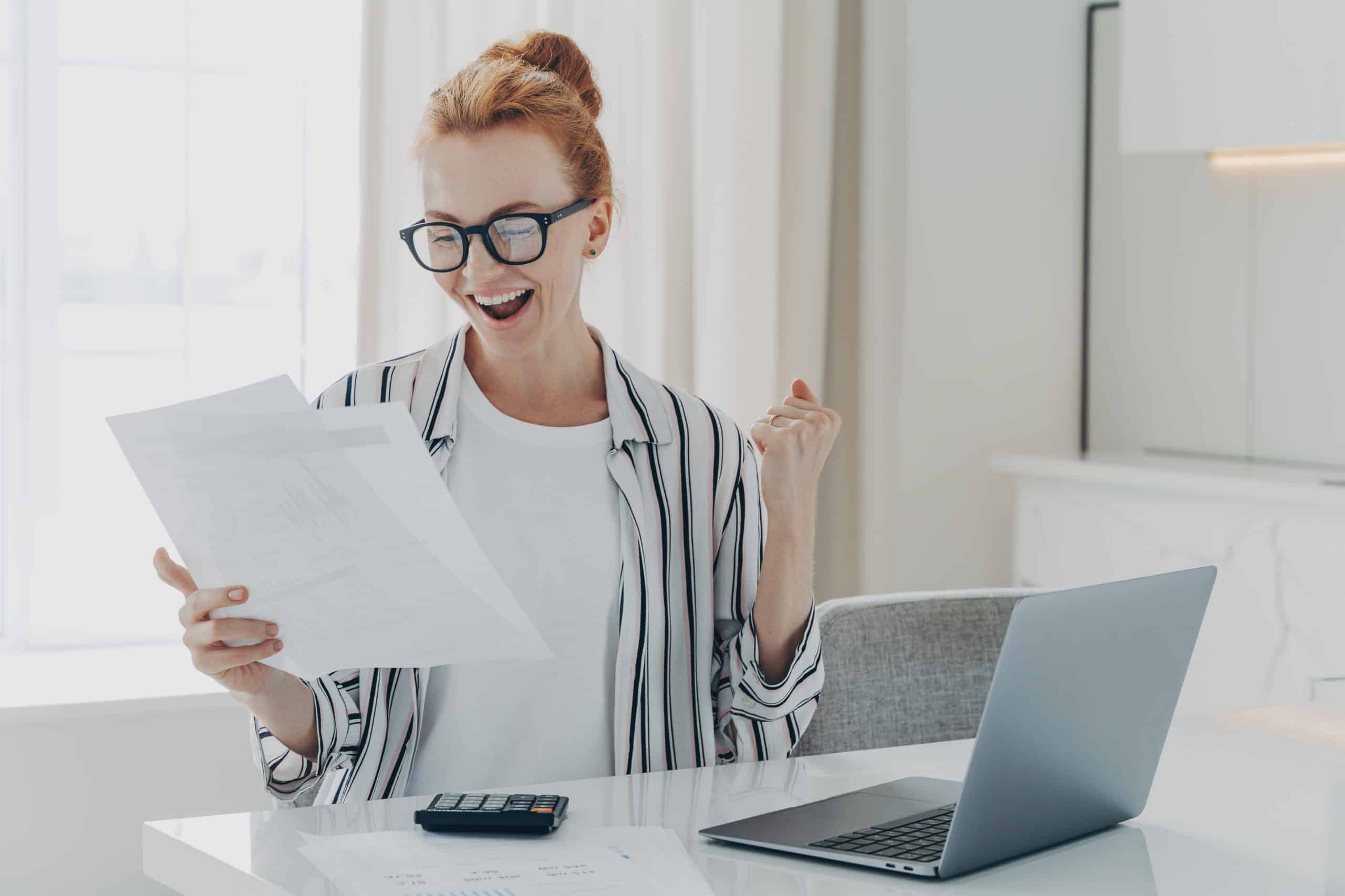 Emotional happy young ginger woman in eyewear holding paper with last mortgage payment banking notification, raising hand with clenched fist, reading document about tax refund from bank
