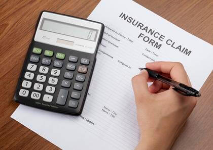 A hand holds a pen filling out an insurance claim form and using a calculator that lays next to the form