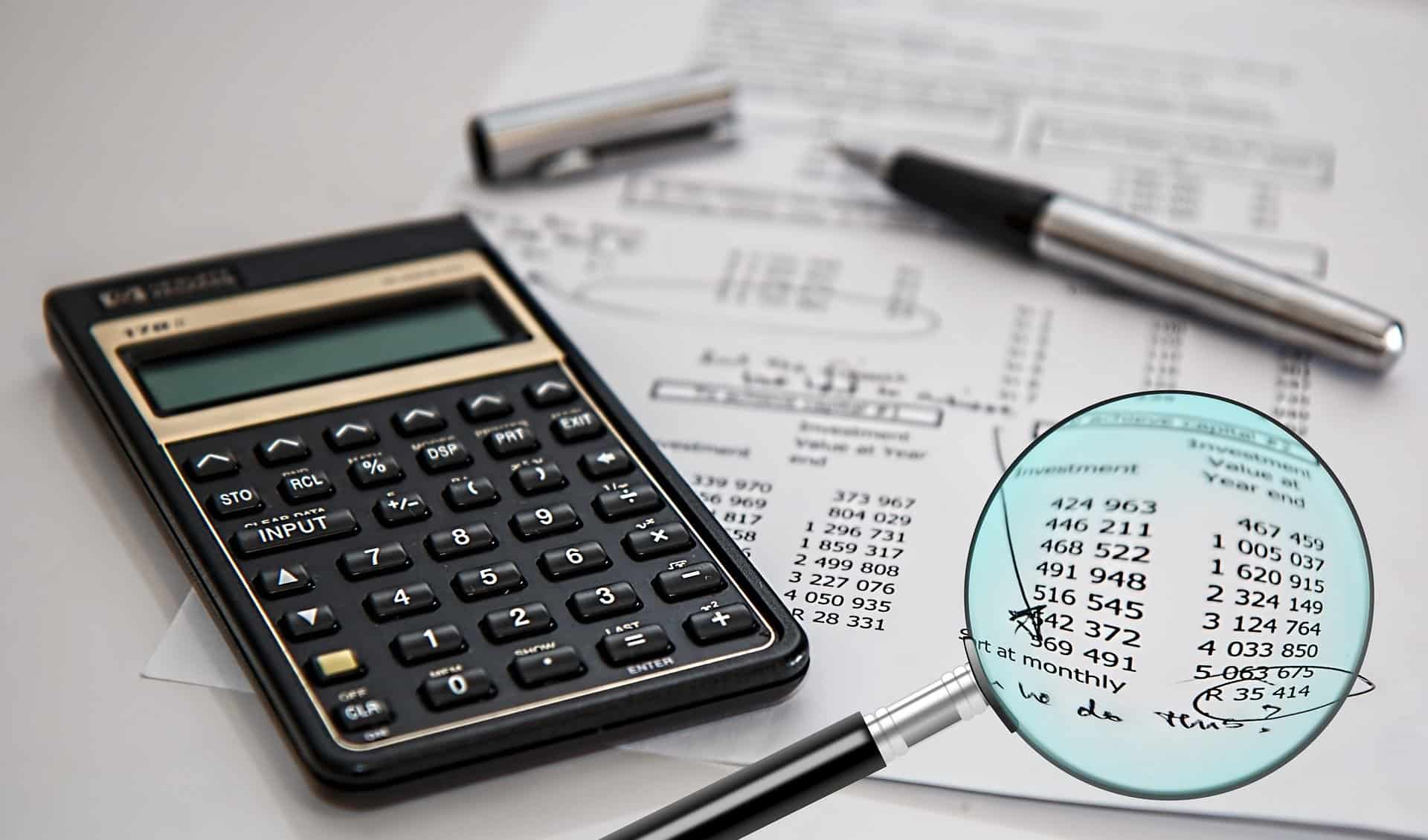 A magnifying glass is used to focus on a section of some bills calculated on a sheet of paper next to an open pen and a calculator