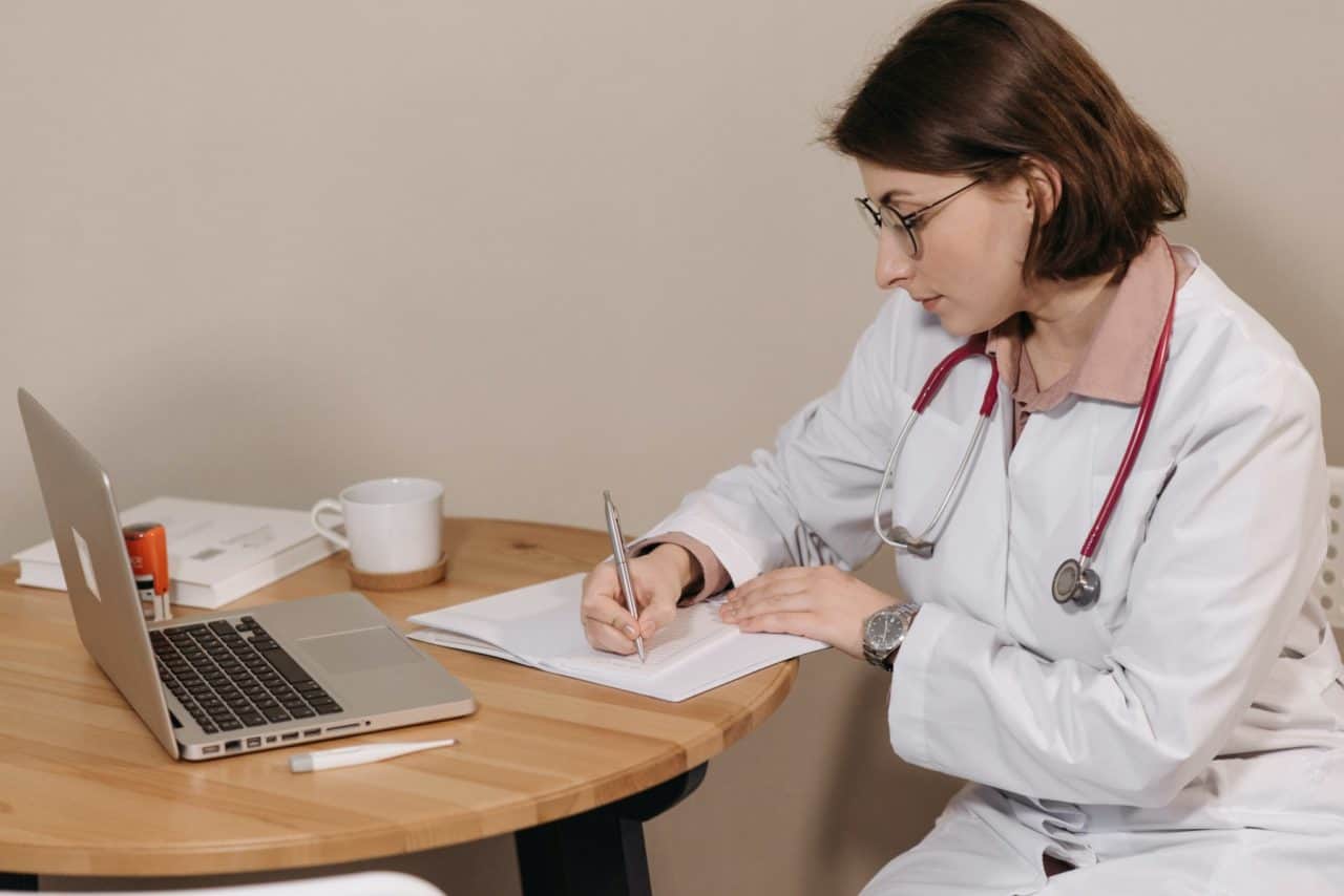 Young doctor sitting at a small table and filling out paperwork next to a laptop, coffee mug, and thermometer