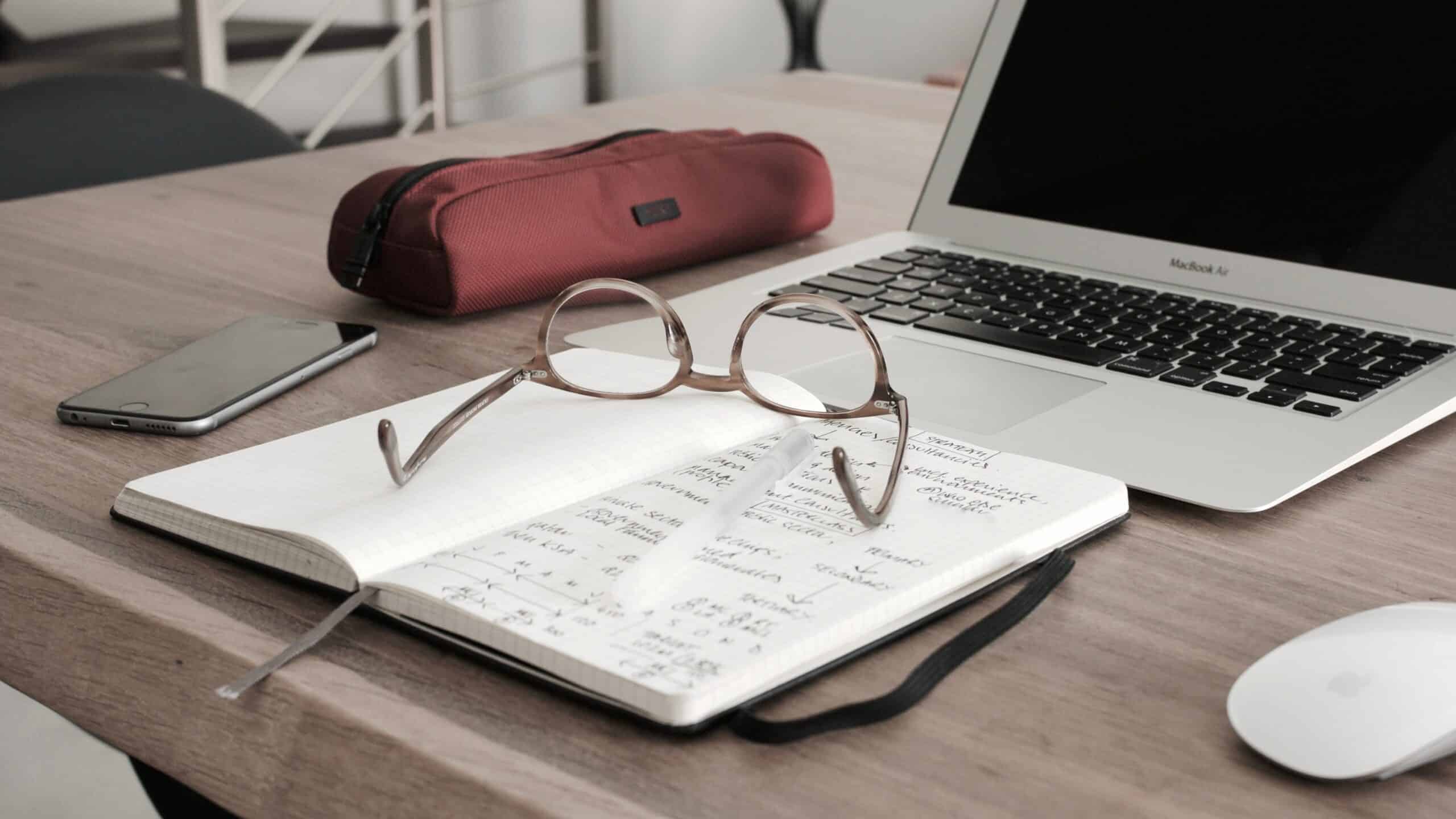Reading glasses holding open a notebook with notes written in it about behavioral health billing, next to a MacBook Air, pencil case, and iPhone