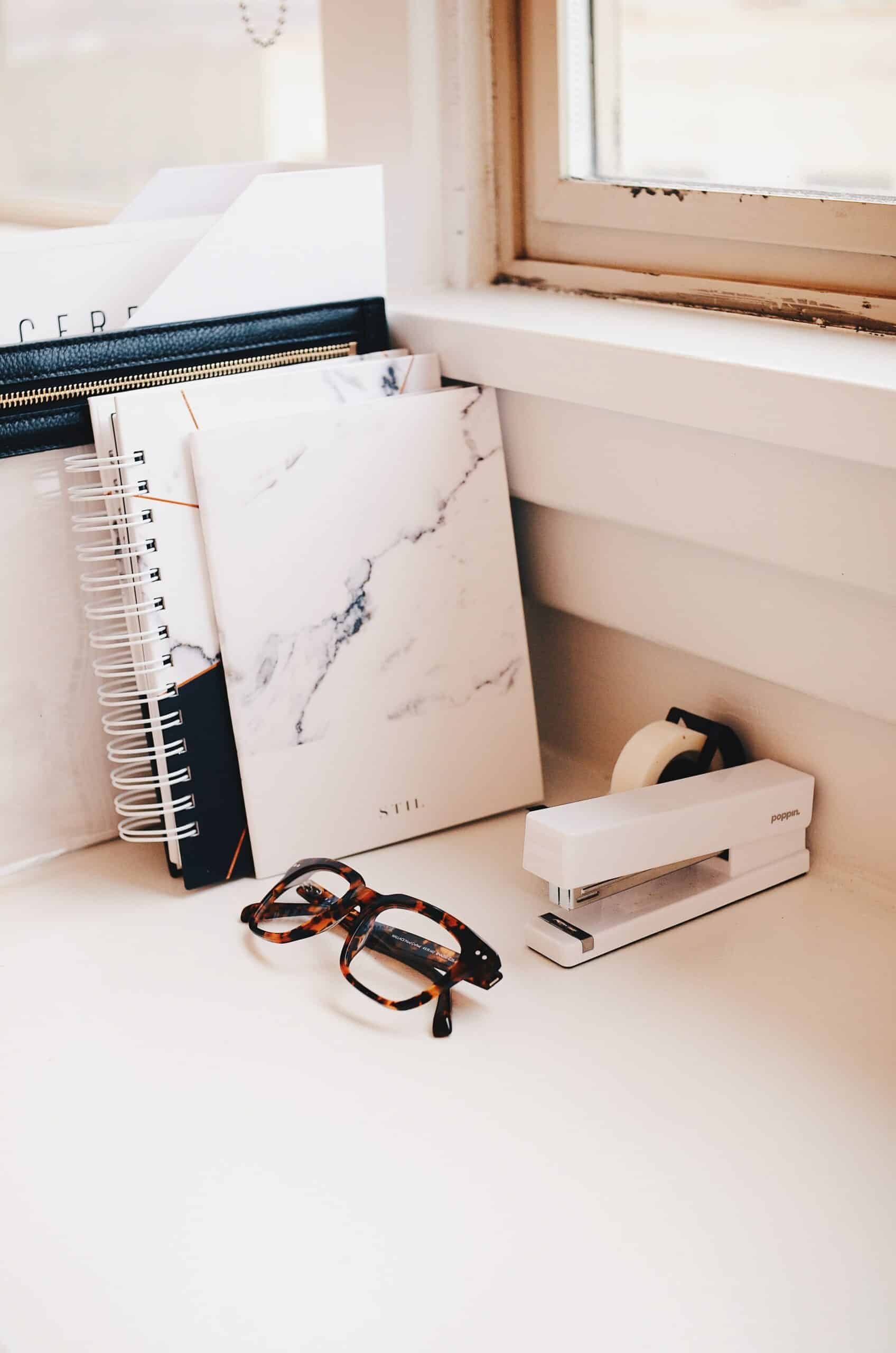 Eyeglasses, notebooks, tape and stapler on a desk.